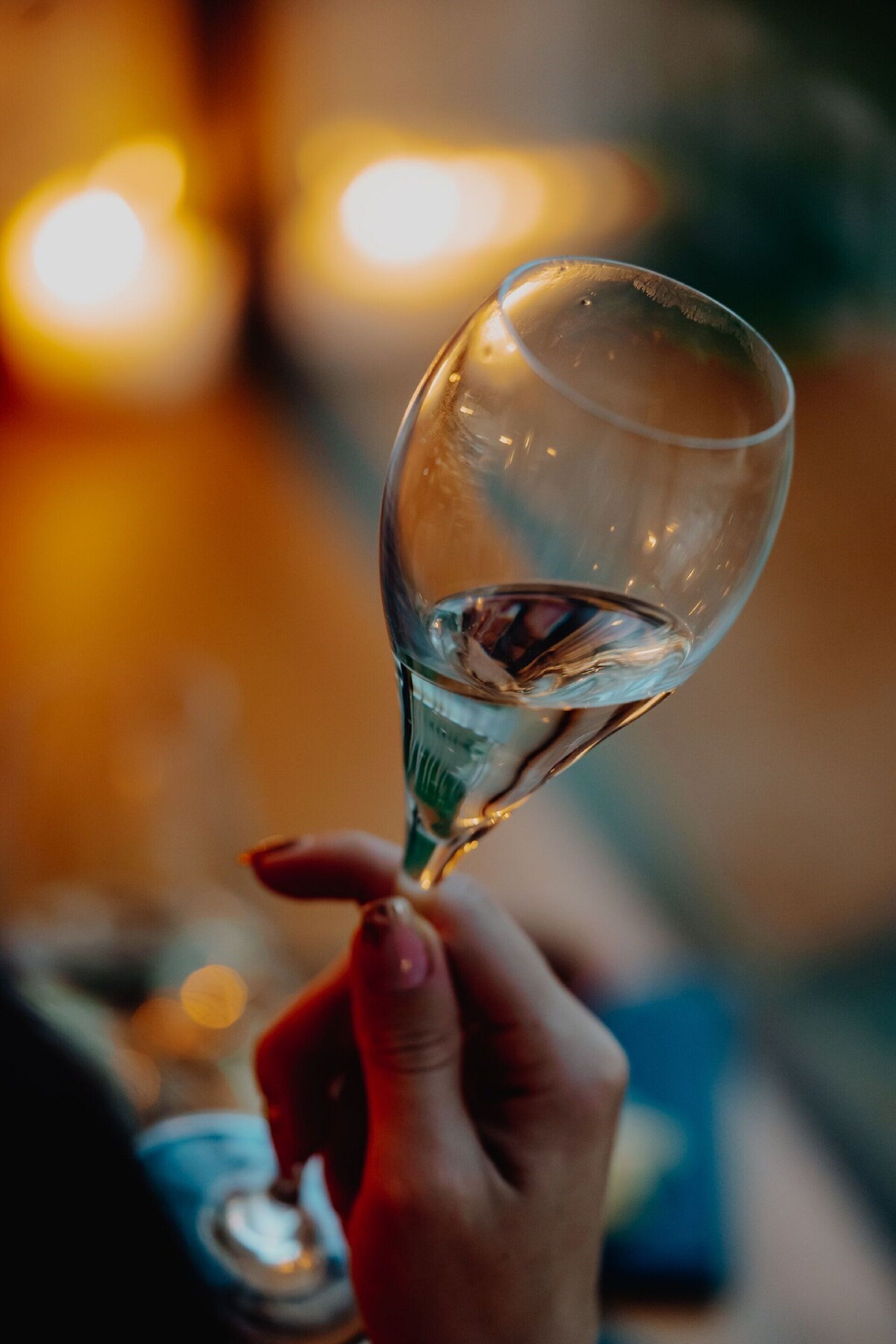 Una mano sostiene un vaso transparente con un líquido, posiblemente una bebida, en un ambiente con poca luz y destellos borrosos de fondo.