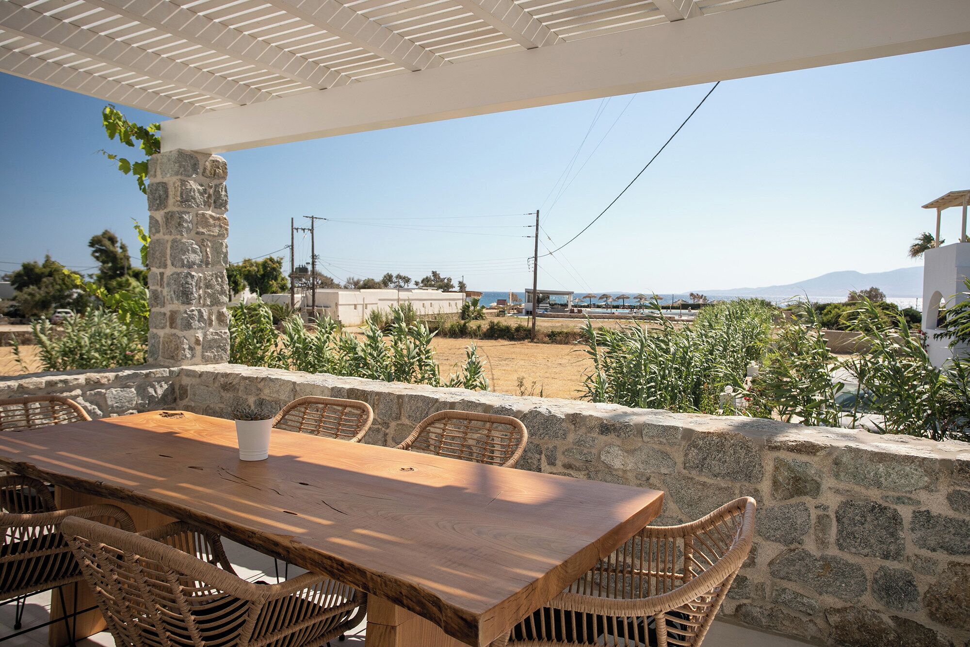 An outdoor patio with a wooden table and wicker chairs, overlooking a coastal area with buildings and a clear sky.