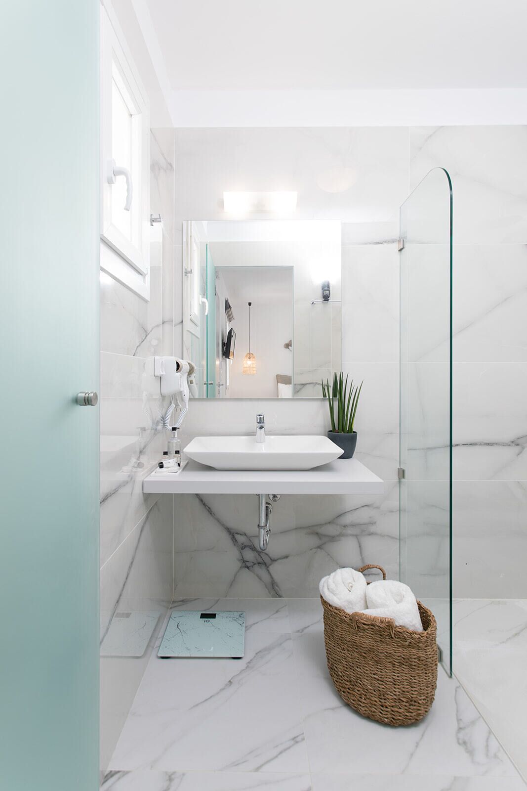 A modern bathroom with a white sink, marble tiles, a glass shower enclosure, and a wicker basket with towels.
