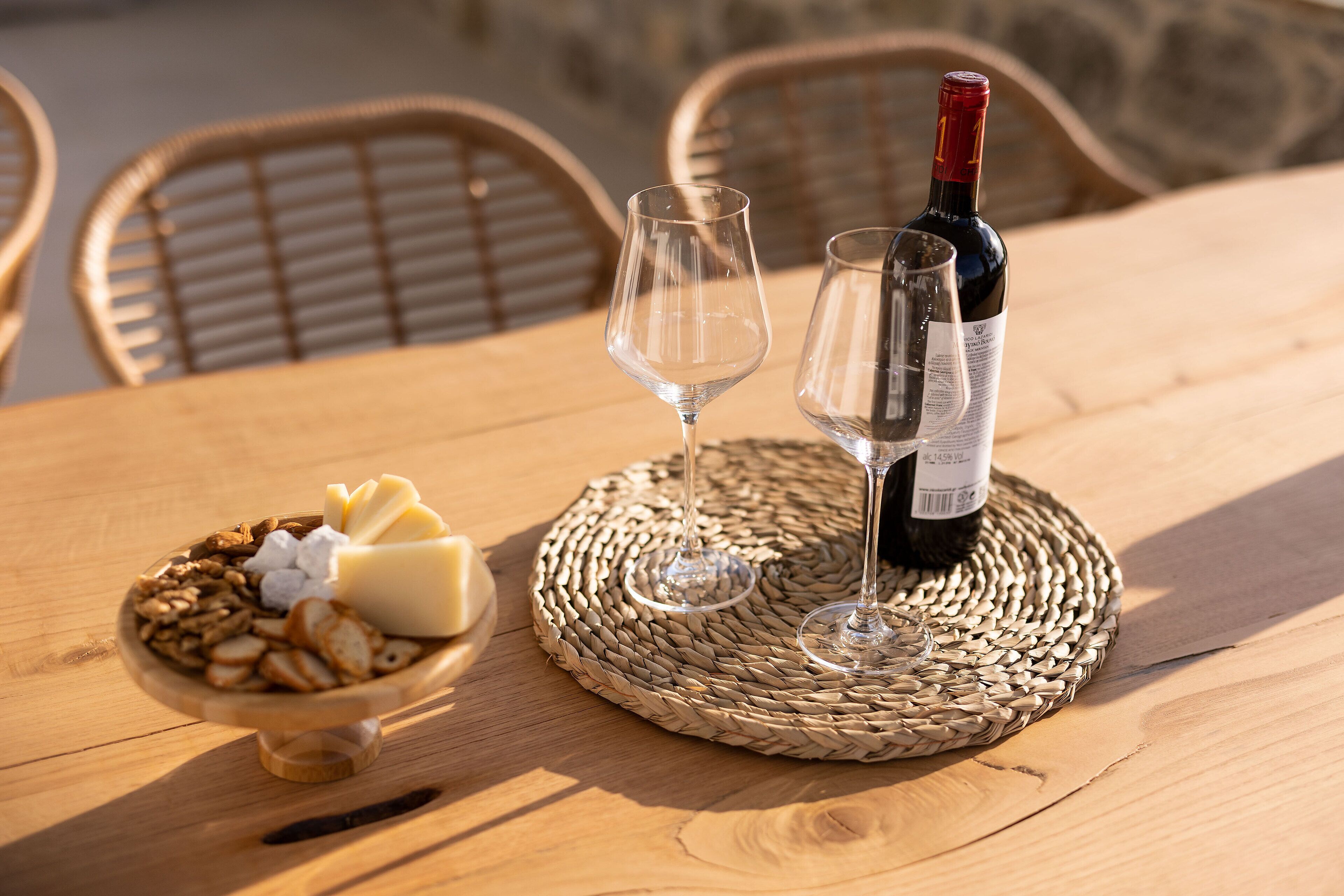 A wooden table with a woven placemat, two wine glasses, a bottle of wine, and a bowl of cheese and crackers.