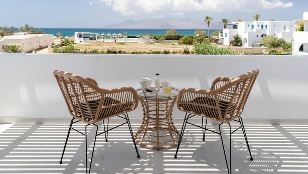 Una terraza con dos sillas de mimbre y una mesa, con vistas a una playa y edificios blancos con techo blanco.