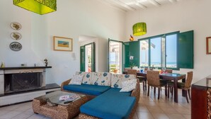 A cozy living room with a sofa, coffee table, and fireplace, leading to a dining area with a view of the sea.