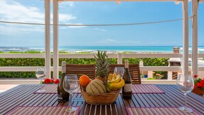 A wooden table with a view of the ocean, a wicker basket with fruits, and two wine bottles.