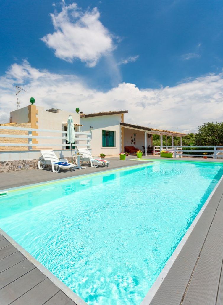 En swimmingpool med liggestole og et overdækket område, omgivet af en bygning med et tagtækket tag og en trappe, der fører til en etage over.