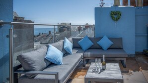 A balcony with a grey sofa and blue cushions, a glass table with a bottle and glasses, and a heart-shaped wreath on the wall. - Ramsey Apartment - 2 Bedroom Apartment - Tenby (Tenby)