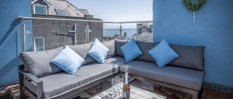 A blue-painted outdoor seating area with a sofa and cushions, a table with a bottle and glasses, and a heart-shaped decoration on the wall.
