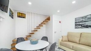 A modern living room with a round table, grey chairs, a beige sofa, a wooden staircase, and two wall-mounted paintings. - Maredimo Village casa 2 (Porto)