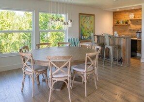 A dining area with a round table and wooden chairs, large windows with greenery outside, a kitchen area with wooden cabinets, and a hanging light fixture. - Spacious 3BR townhome w/ easy beach & pool access (Siesta Key)