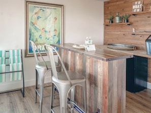 A bar area with a wooden counter, two metal bar stools, a poster on the wall, and a wooden shelf with items.