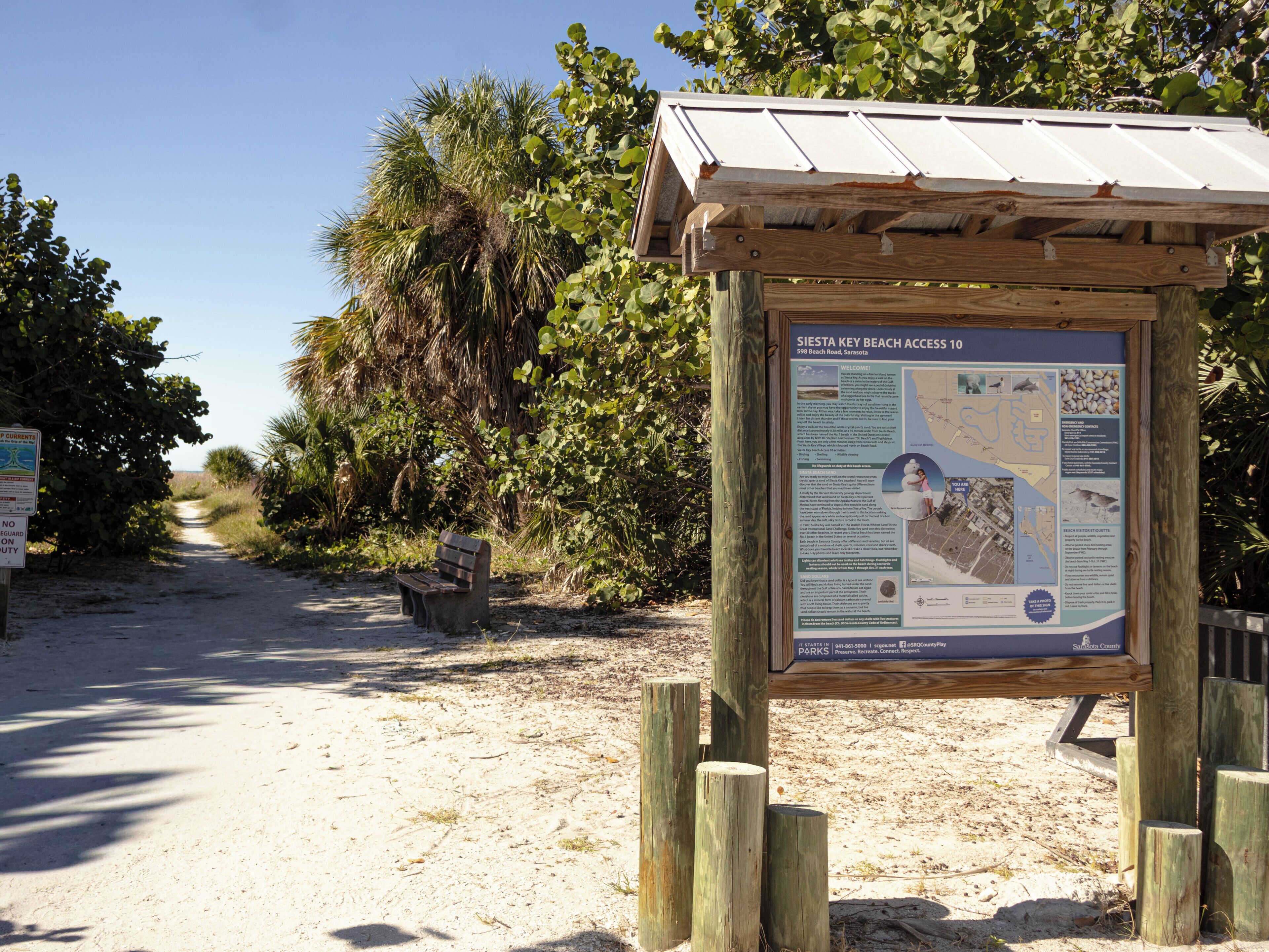 A wooden information board with text and maps, a bench, and a sandy path leading to a beach.