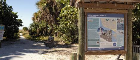 A wooden information board with text and maps, a bench, and a sandy path leading to a beach.