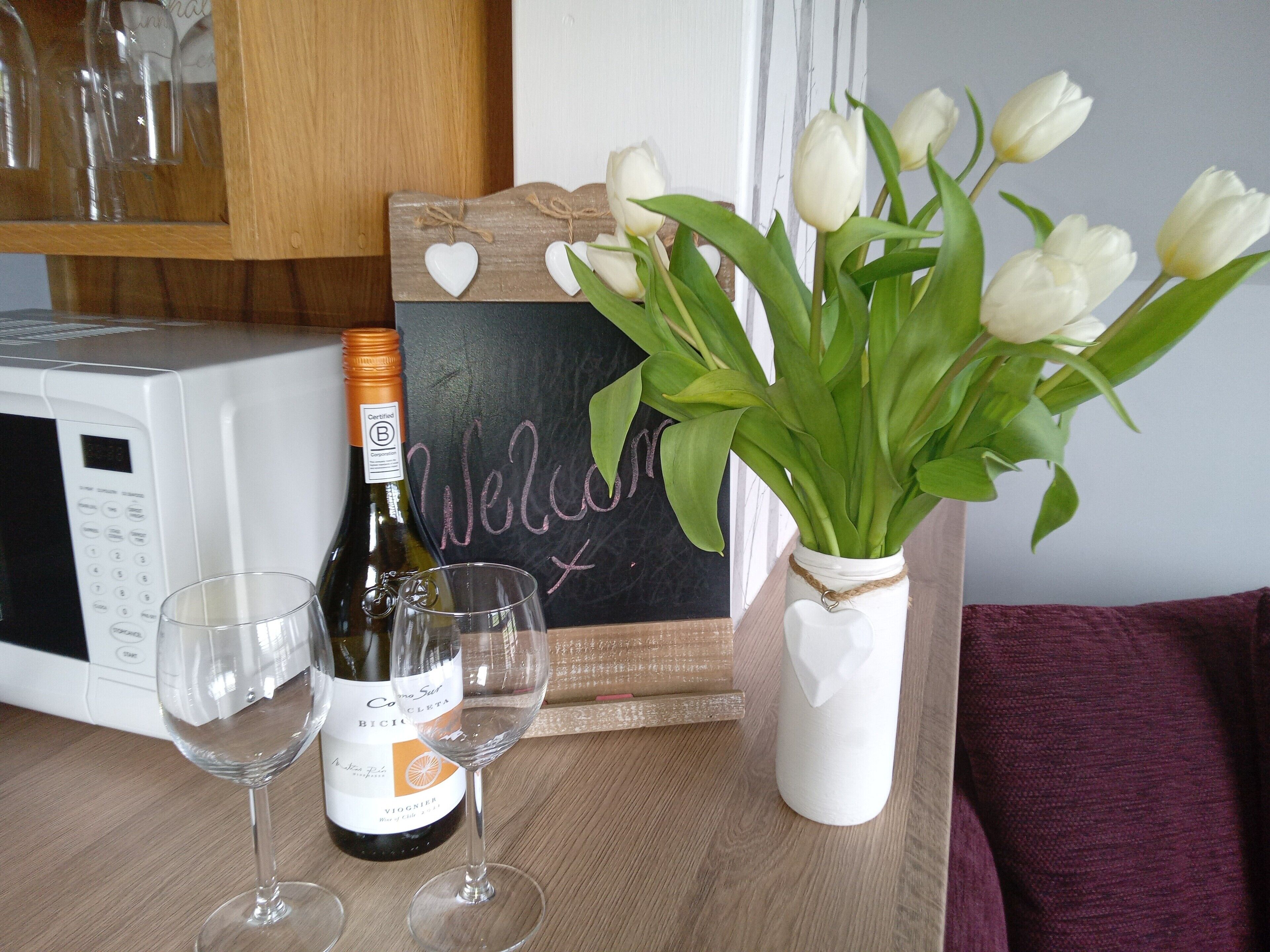 Ein Tisch mit einer Weinflasche, zwei Gläsern, einer Vase mit weißen Tulpen und einer Tafel mit der Aufschrift 'Welcome'.