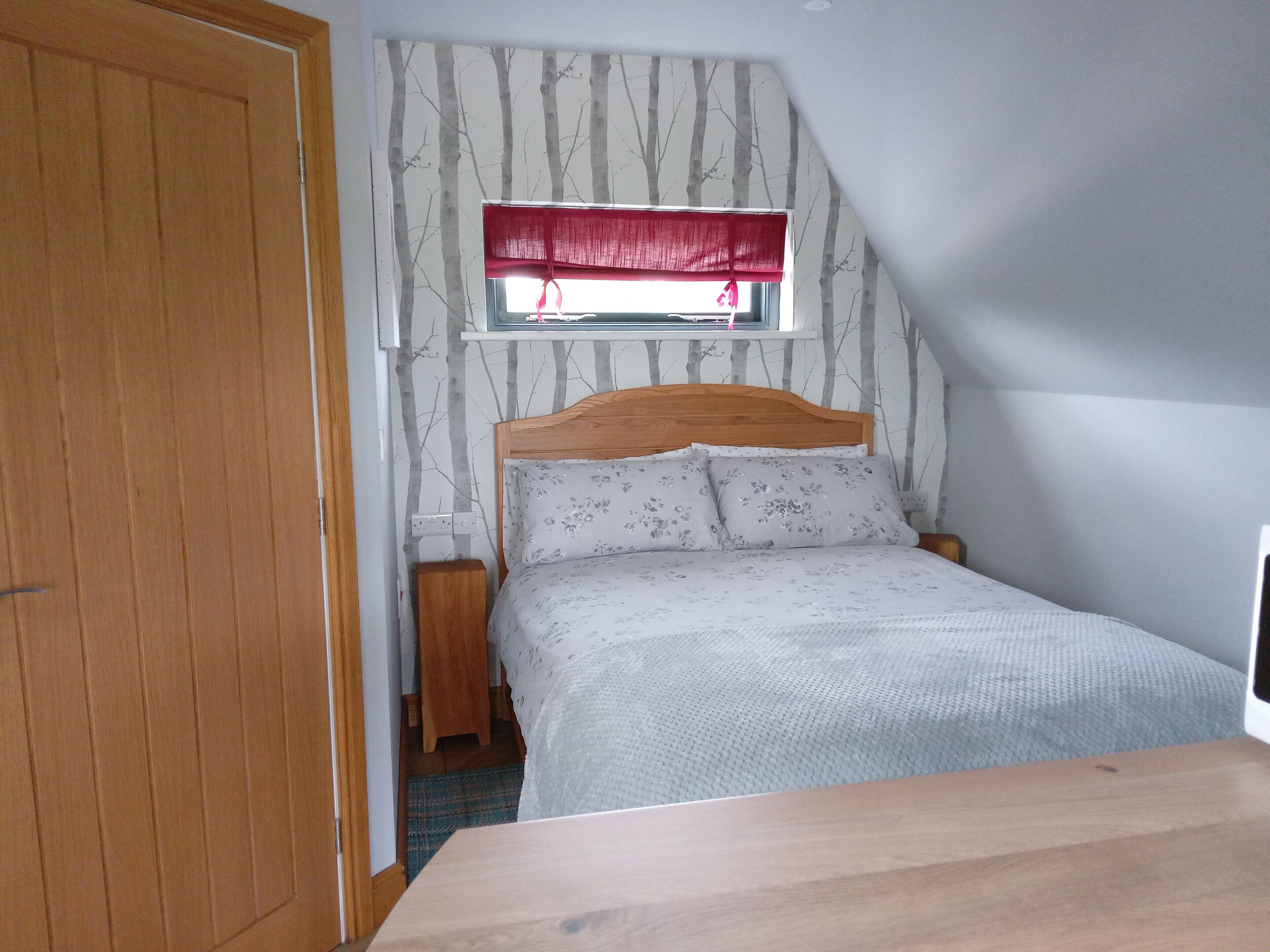 Een slaapkamer met een houten hoofdbord, een bed met bloemenbeddengoed, een raam met een rode vouwsluier en een houten nachtkastje.