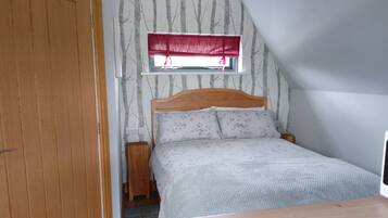 Een slaapkamer met een houten hoofdbord, een bed met bloemenbeddengoed, een raam met een rode vouwsluier en een houten nachtkastje.