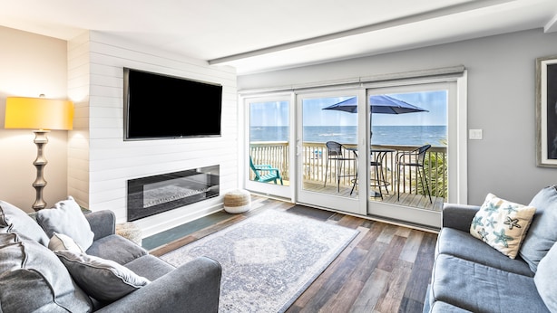A living room with a fireplace, a flat-screen TV, a sofa, and a view of the ocean through large sliding glass doors. - Seagrove Villa 11C - Extreme Oceanfront! Resort! (Isle of Palms)