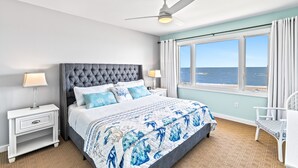 A bedroom with a large bed, a ceiling fan, a nightstand with a lamp, a chair, and a view of the ocean through a window. - Seagrove Villa 11C - Extreme Oceanfront! Resort! (Isle of Palms)