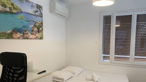 Een slaapkamer met een bed, een bureau, een stoel, een schilderij, een airconditioner en een raam met jaloezieën.