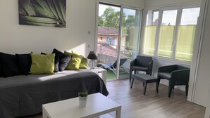 Een moderne woonkamer met een bank, een salontafel en een stoel. Er is een balkon met uitzicht op gebouwen en bomen.