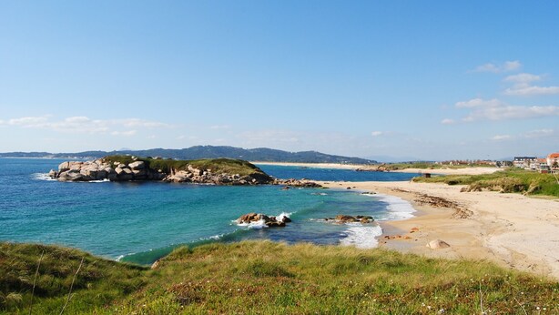 Eine Küstenlandschaft mit einem Felsvorsprung, einem Sandstrand und sanften Hügeln im Hintergrund.