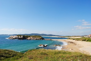 A coastal landscape with a rocky outcrop, sandy beach, and distant hills.