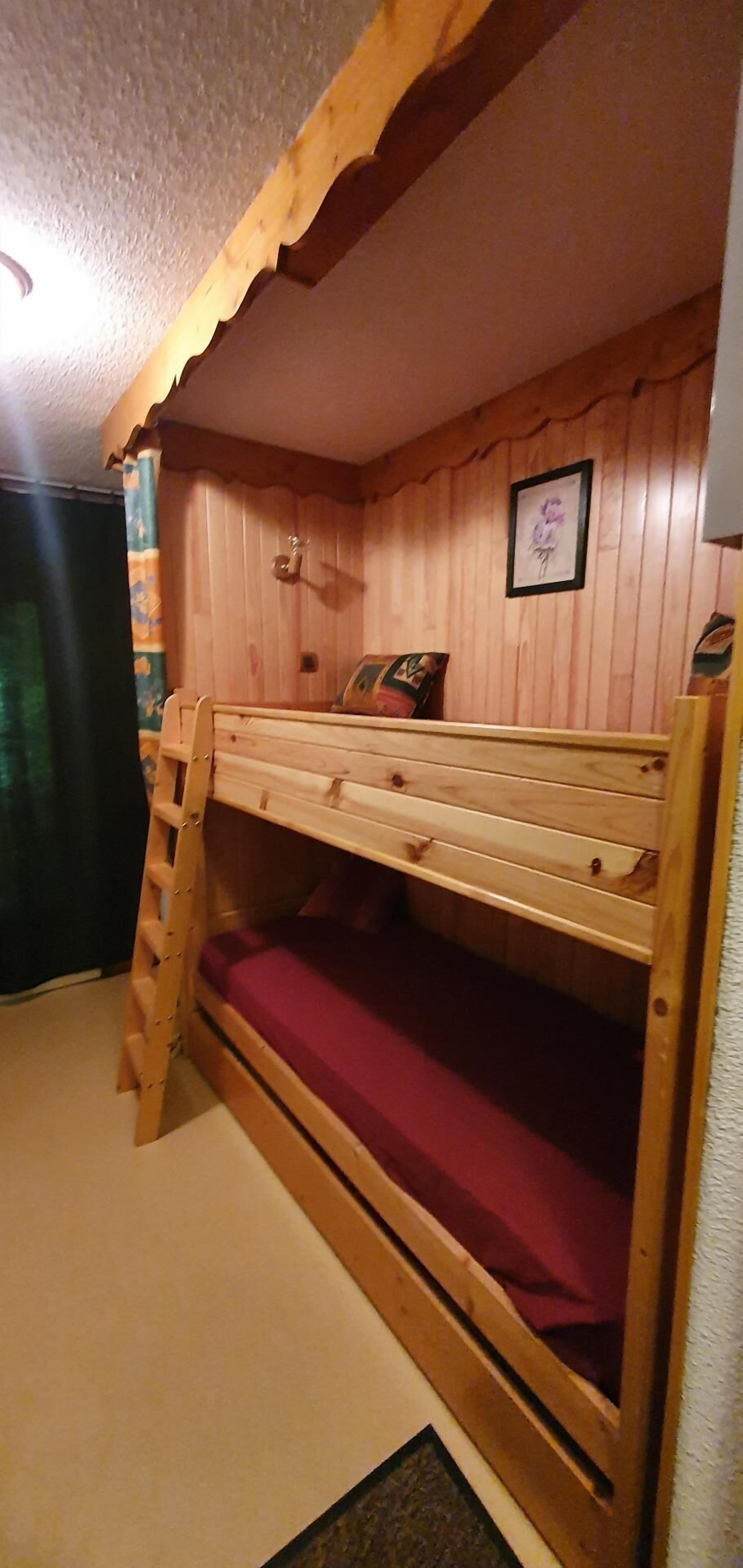 Ein Etagenbett mit Holzrahmen und einer roten Matratze. Eine Leiter befindet sich auf der linken Seite. Der Raum hat Holzwände und ein gerahmtes Bild an der Wand.