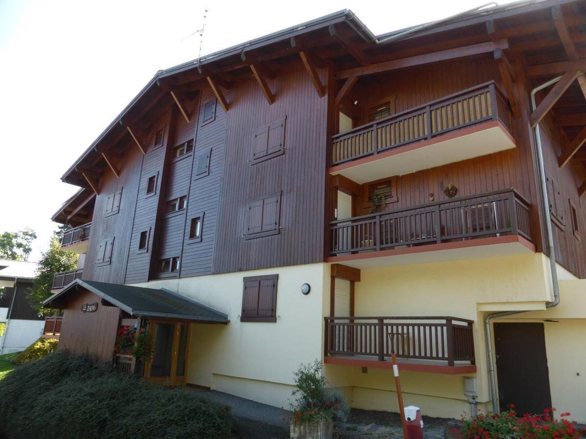 A multi-story wooden building with balconies, a small entrance, and a garden area with flowers.