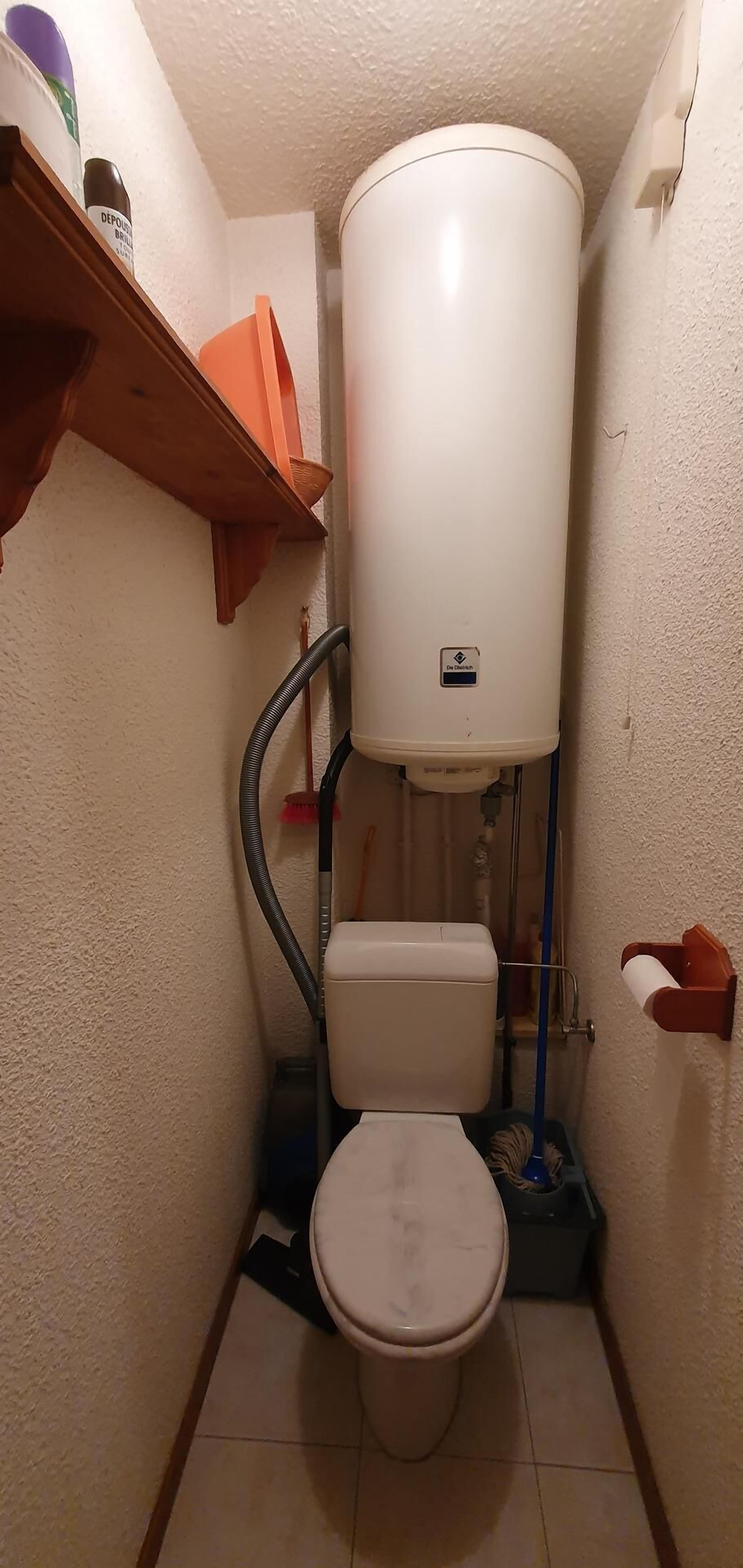 A small bathroom with a toilet, a white water heater, and a wooden shelf.