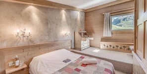 Um quarto com cabeceira de madeira, mesa de cabeceira com garrafa e copo, cama com colcha, cômoda e janela com vista para as montanhas.
