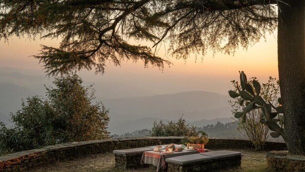 A rustic outdoor seating area with a table set for a meal, surrounded by natural vegetation and overlooking a mountainous landscape. - Mary Budden Estate (Almora)