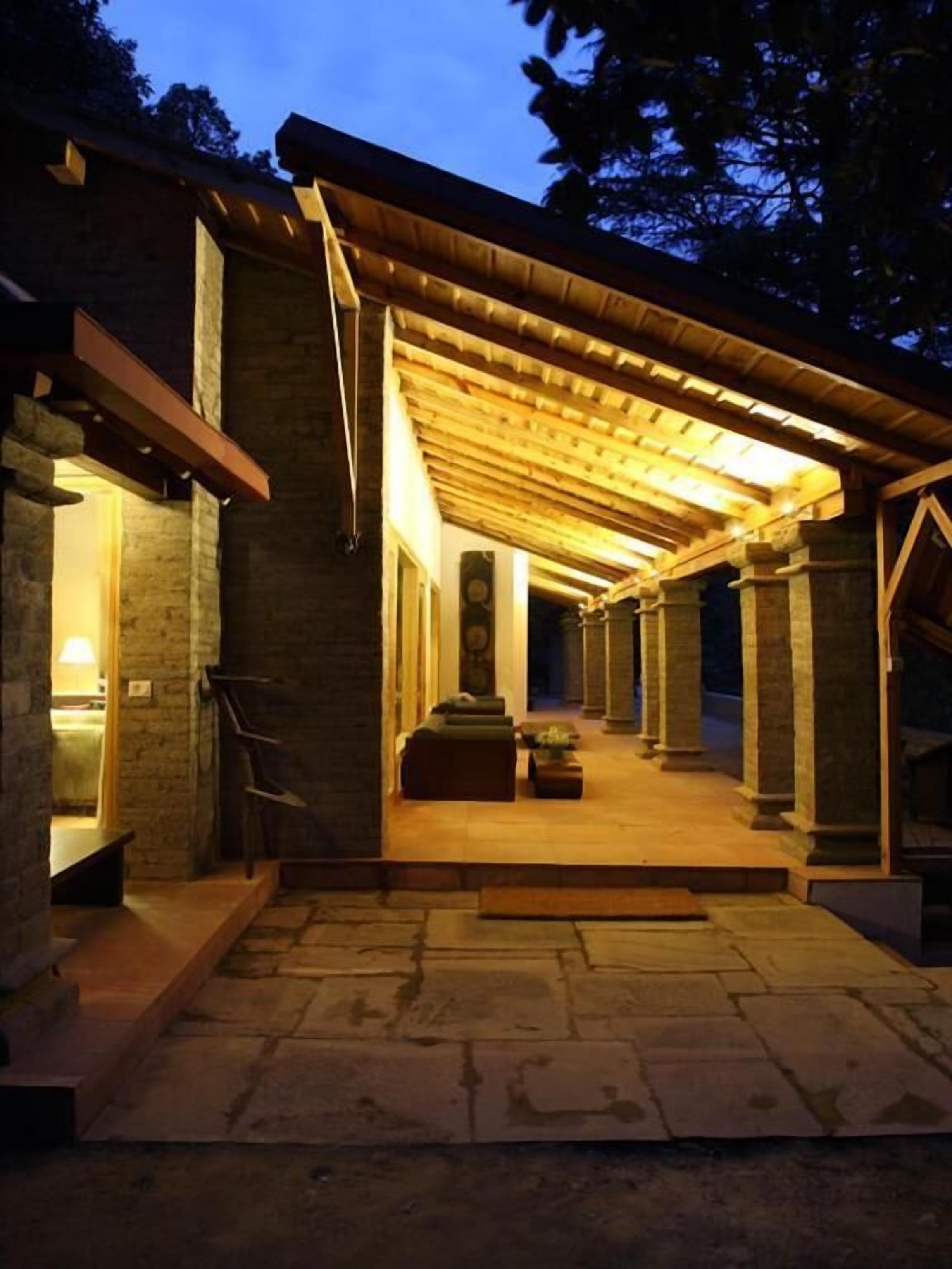 Un espace extérieur abrité, doté de murs en pierre et de poutres en bois, qui mène à un espace intérieur aménagé avec des sièges et un escalier.