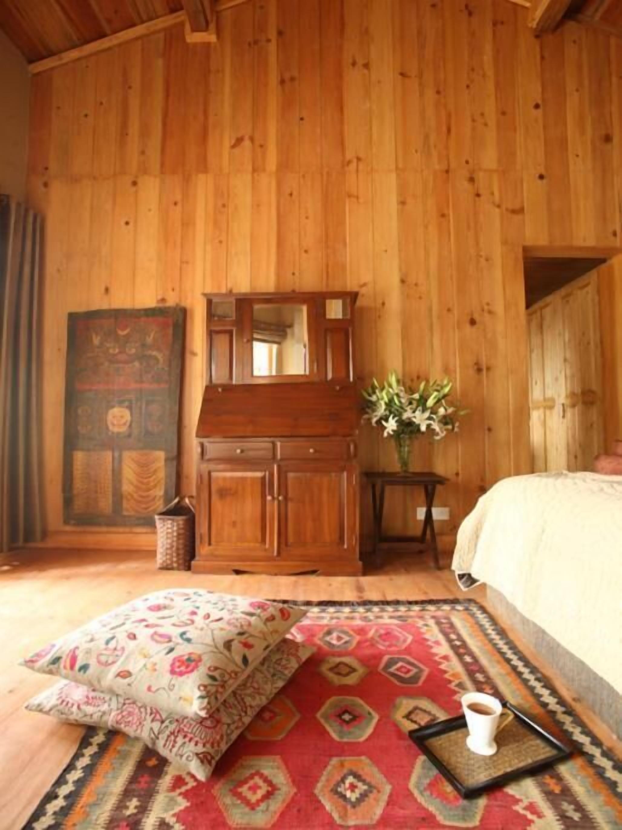 A wooden-paneled room with a bed, a dresser, a mirror, a vase with flowers, and a patterned rug.