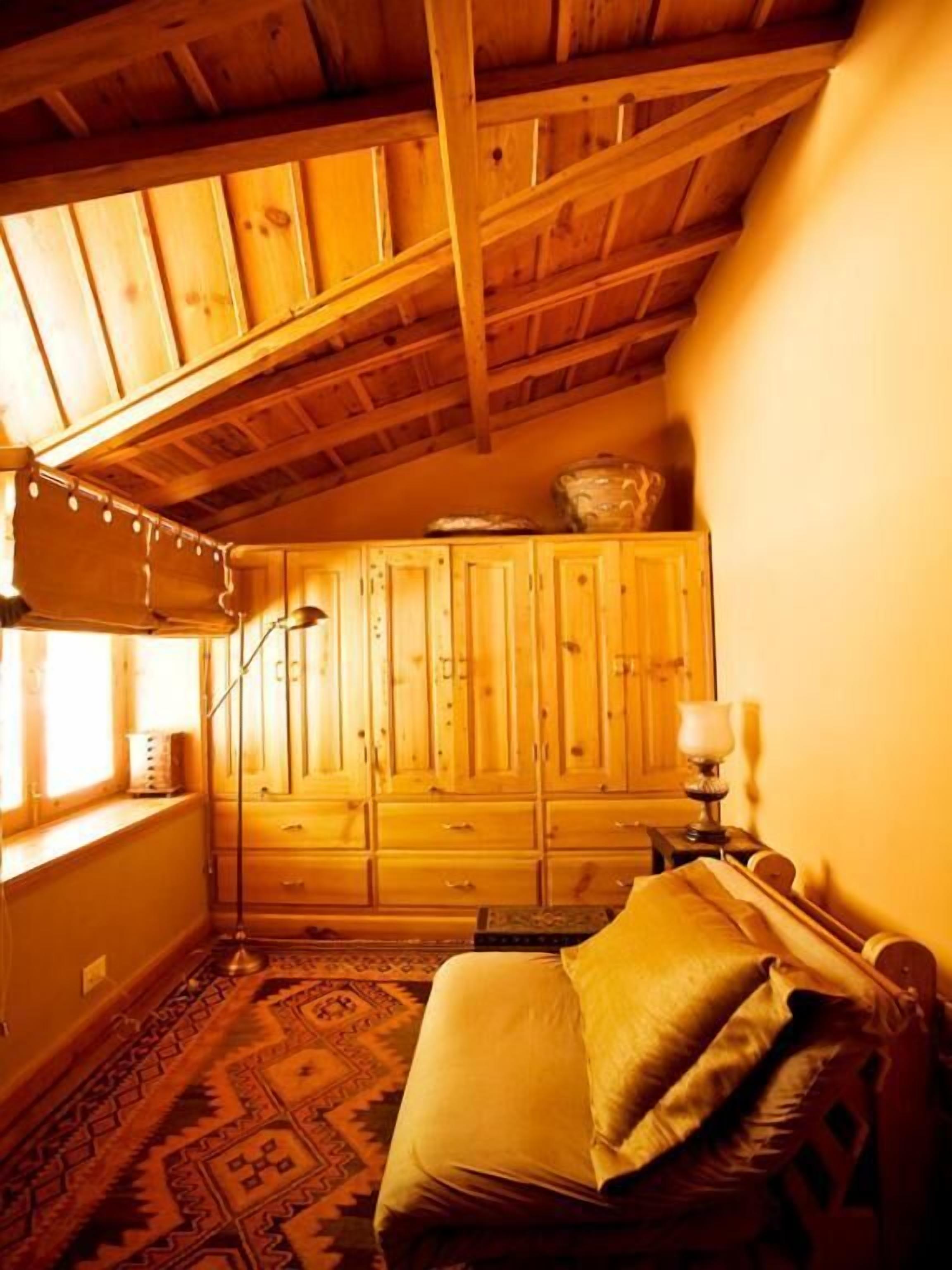 Ein gemütliches Zimmer mit Holzbalkendecke, einem Bett mit Kissen, einer Lampe und einem Fenster mit Vorhängen.