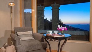 A cozy room with a grey armchair, a small round table with a vase of flowers, and a view of mountains through a large window. - Mary Budden Estate (Almora)