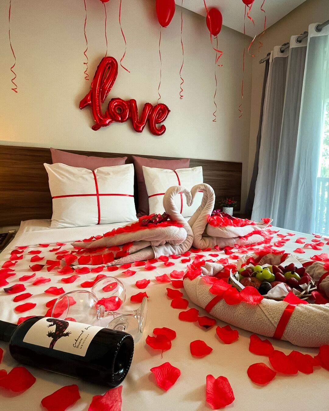 A hotel room with a bed adorned with rose petals, a bottle of wine, and heart-shaped towels.