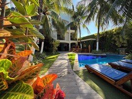 Área da piscina com espreguiçadeiras, piscina cristalina e jardim bem cuidado com plantas tropicais.