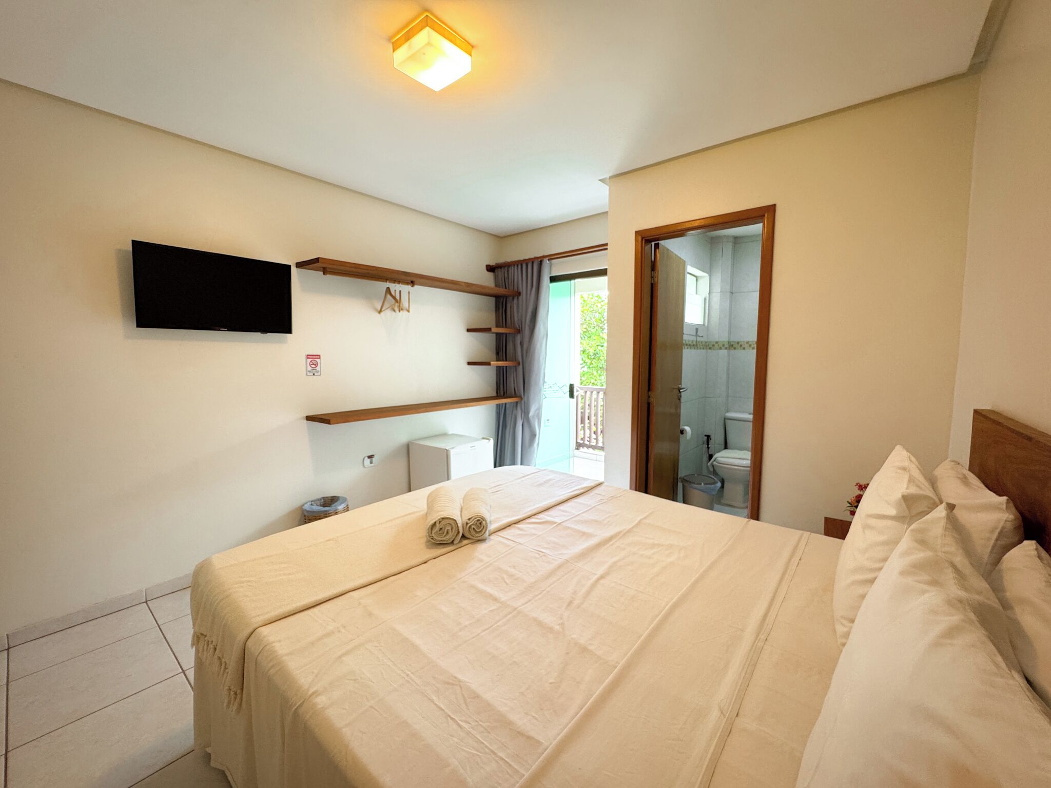 Un dormitorio con cama, televisor de pantalla plana, mesita de noche pequeña, estante en la pared y puerta que da al baño.