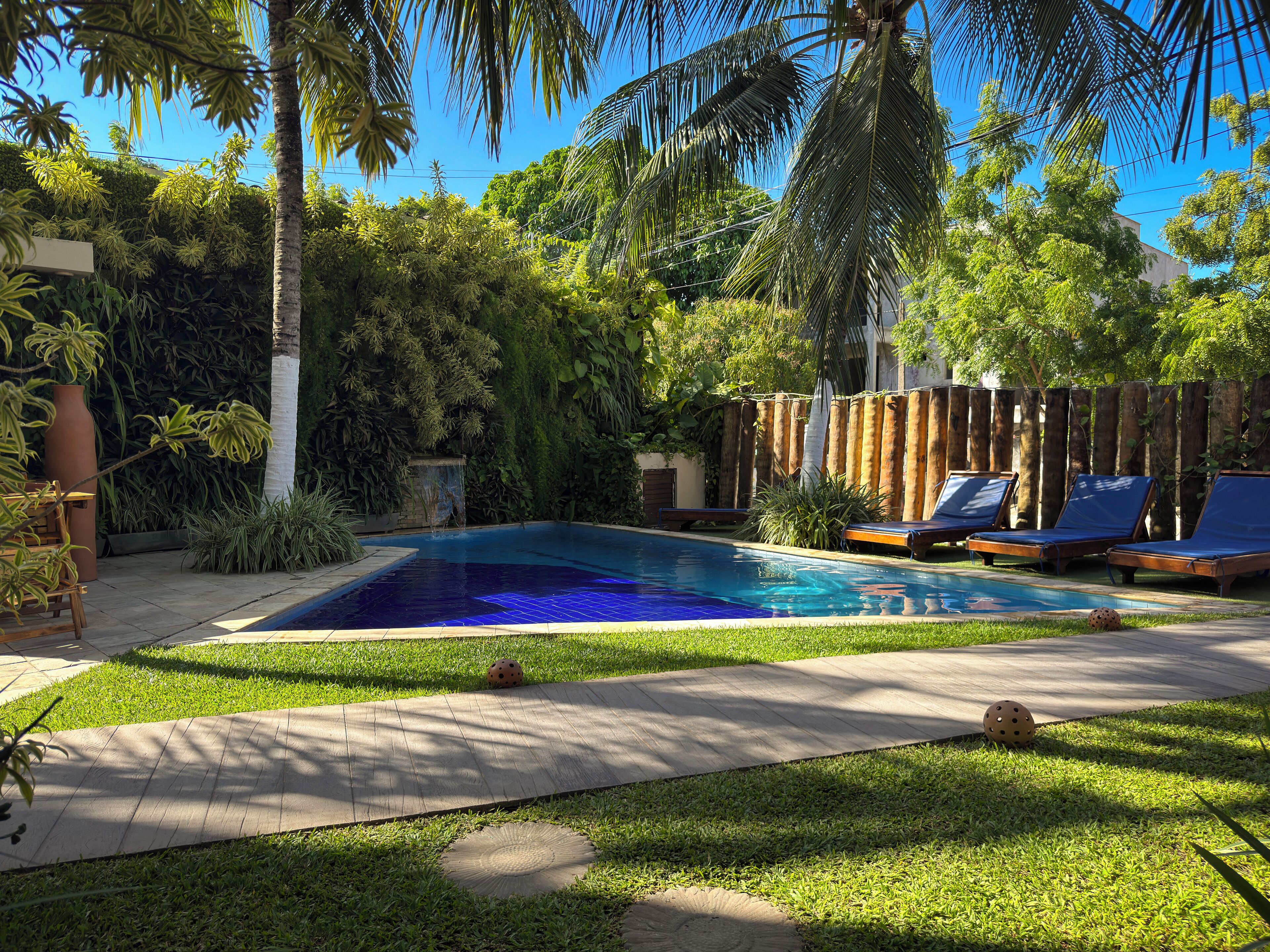Ein rechteckiger Swimmingpool mit umlaufender Holzterrasse, einem Holenzaun und mehreren Liegestühlen.