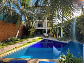 Una piscina con solárium de baldosas, rodeada de palmeras y un jardín cuidado.