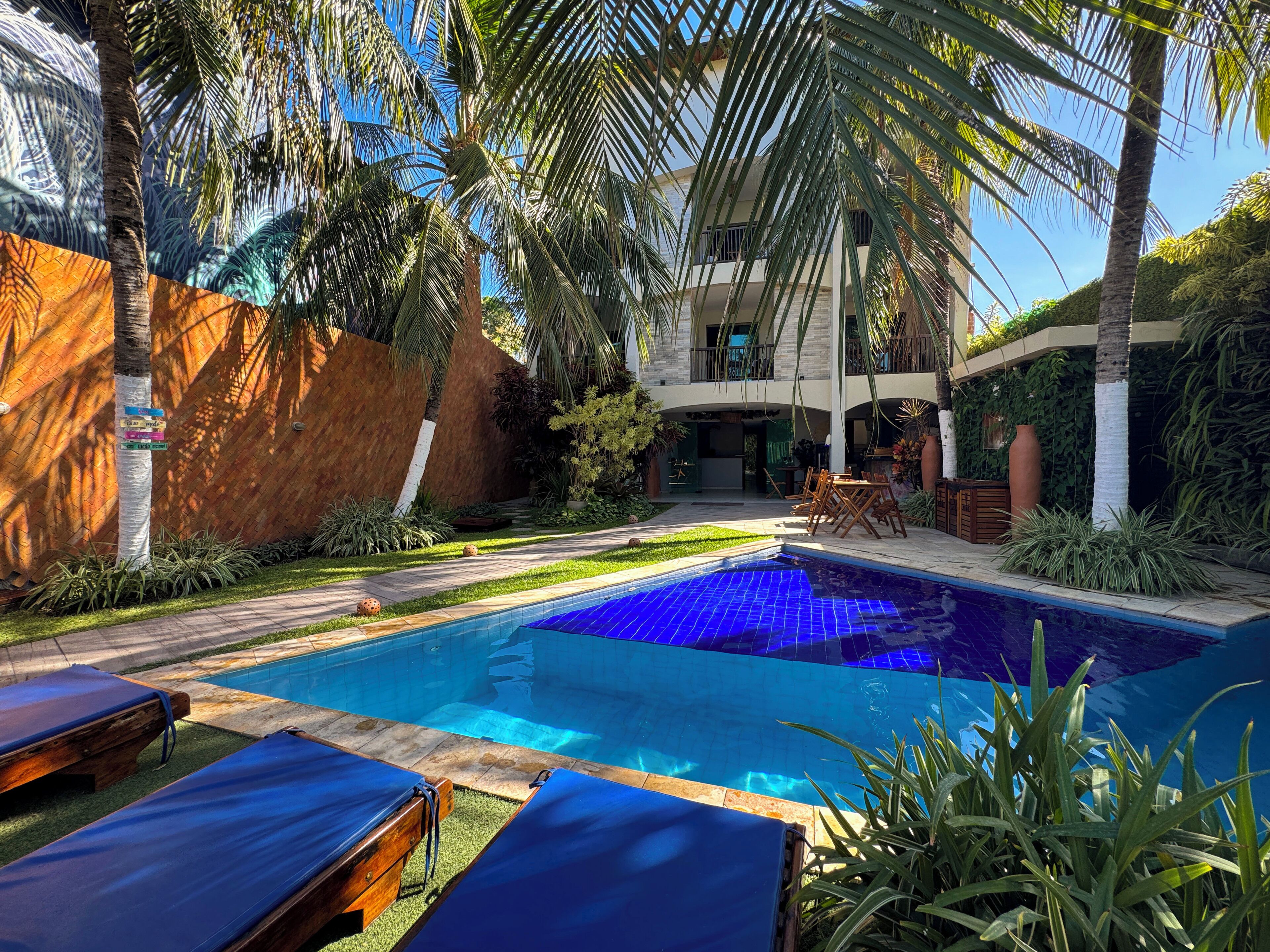 Zona de piscina con sillones reclinables, palmeras y un edificio de dos pisos con balcón.
