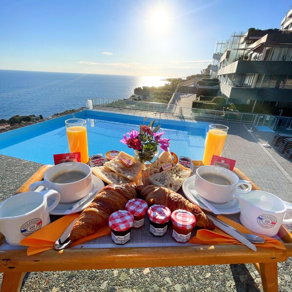 A breakfast tray with croissants, pastries, and jams on a balcony overlooking a pool and the sea.