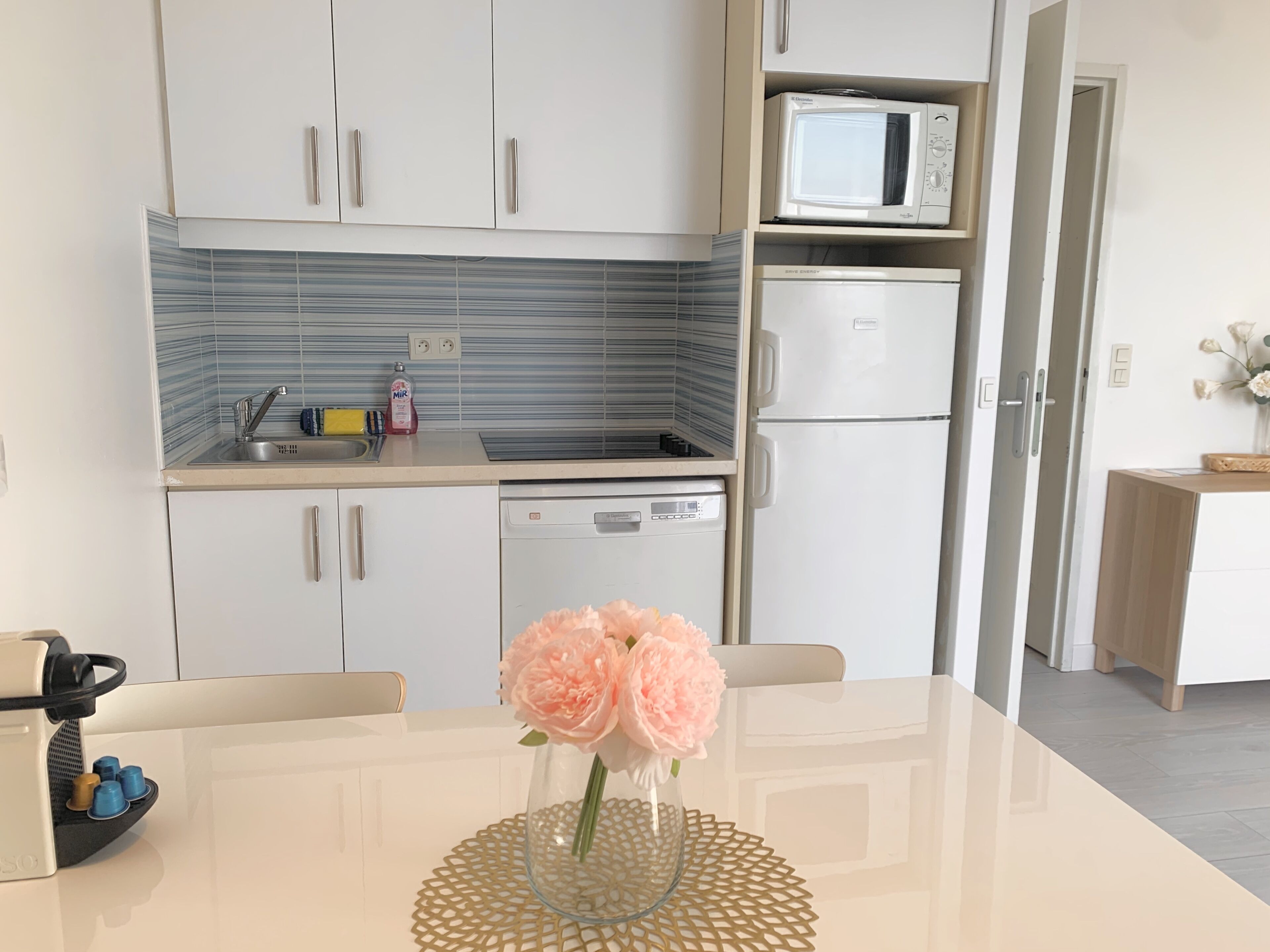 Eine moderne Küche mit weißen Schränken, einer Mikrowelle, einem Kühlschrank und einem Geschirrspüler. Ein Tisch mit einem Blumenstrauß steht daneben.