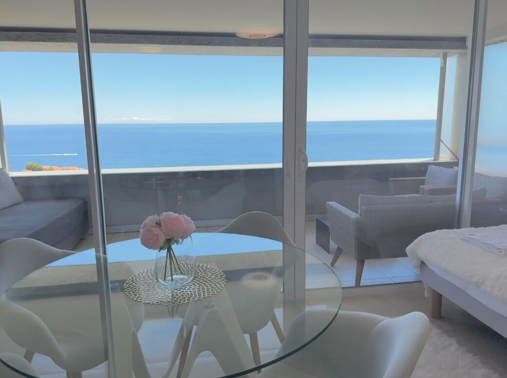 A modern room with a glass table and chairs, a bed, and a view of the sea.
