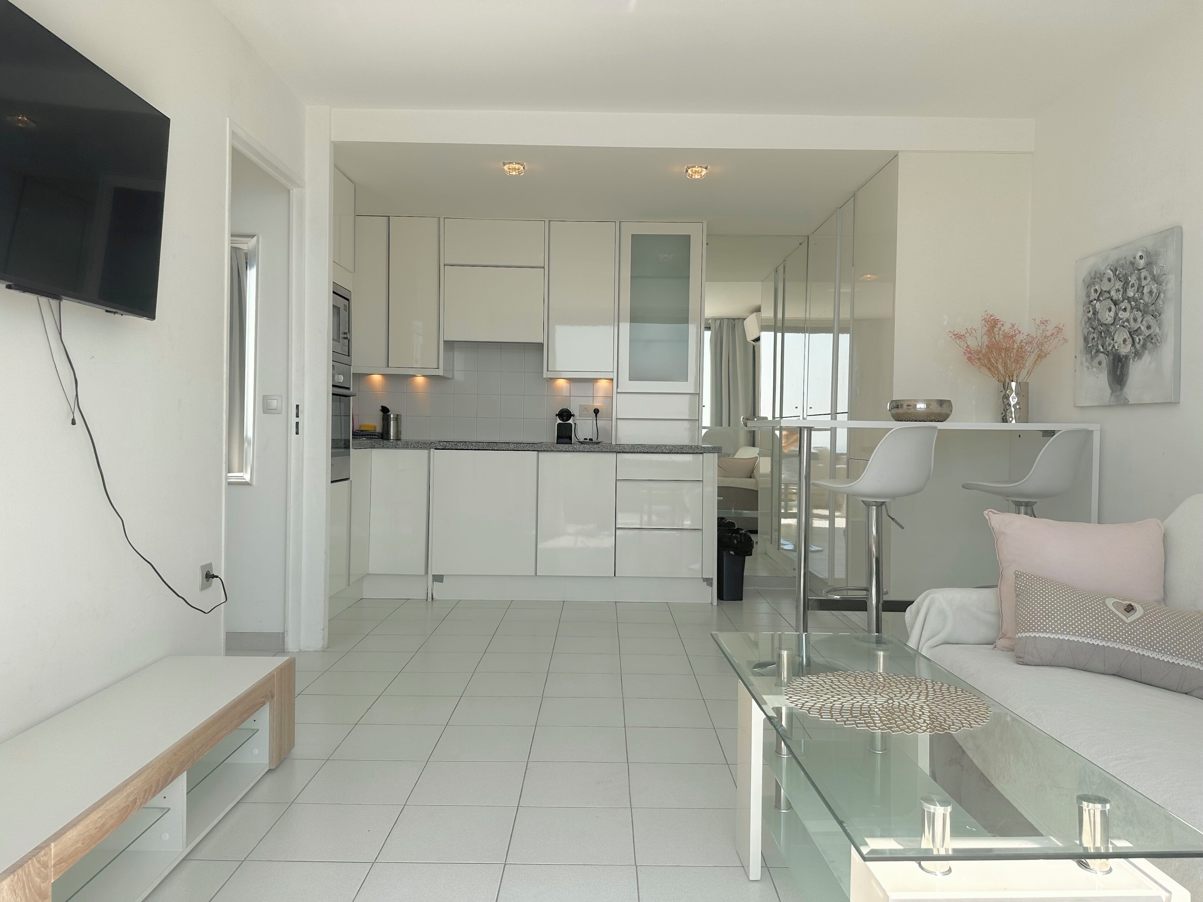 A modern kitchen with white cabinets, a dining area with a glass table, and a living room with a sofa and a coffee table.