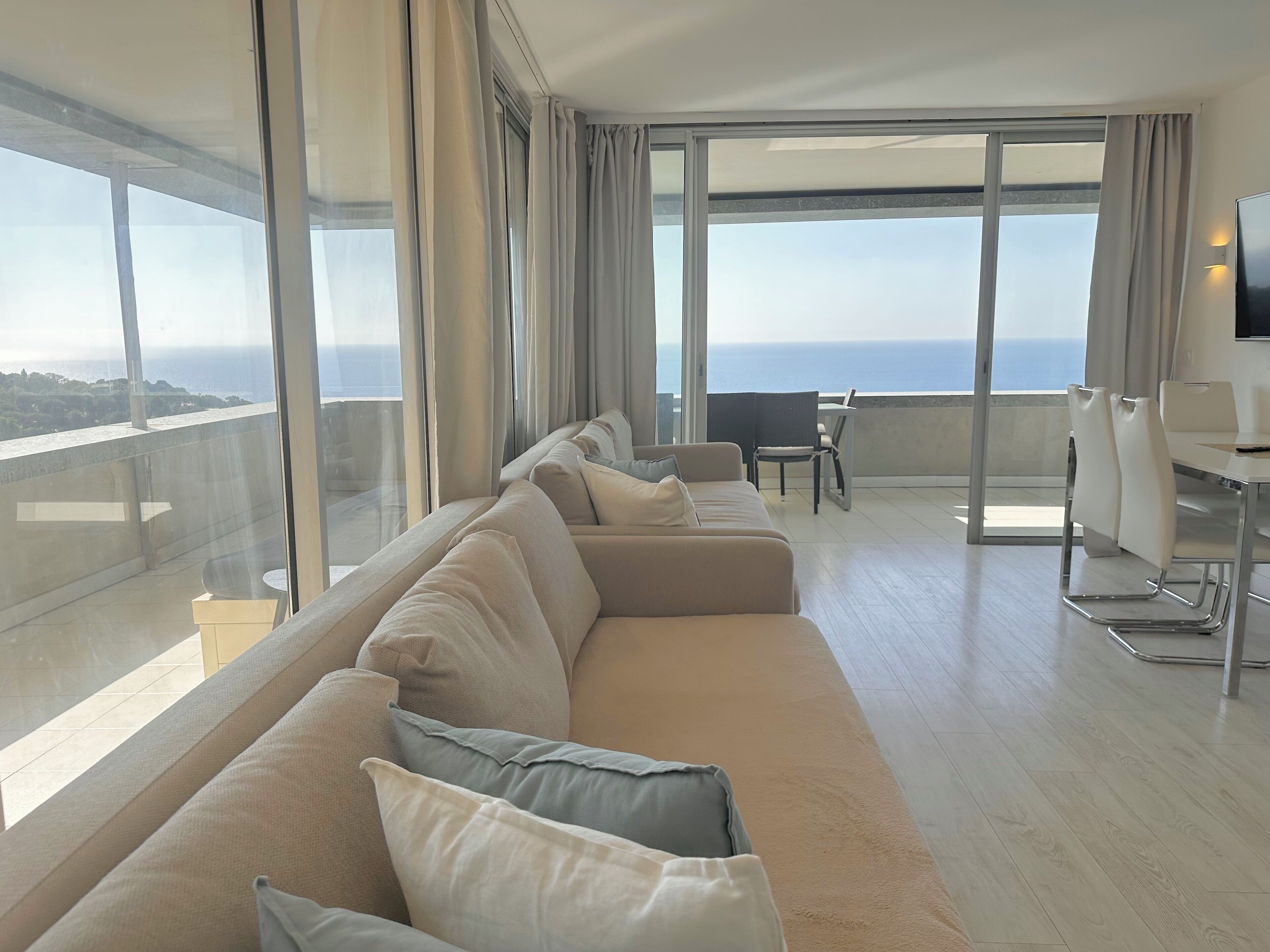 A modern living room with a long beige sofa, a dining table with chairs, and large windows offering a view of the sea.