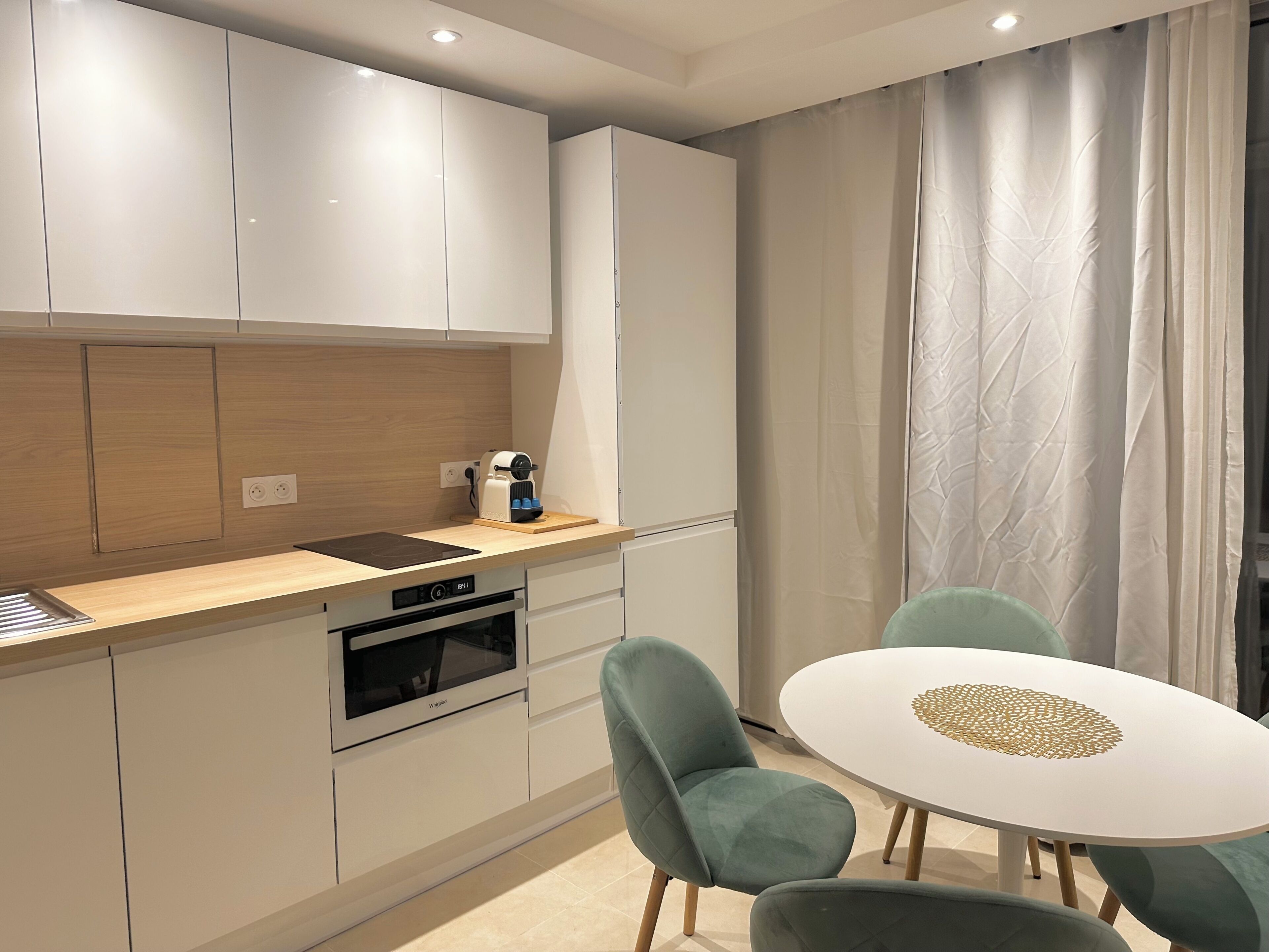A modern kitchen with white cabinets, a built-in oven, and a coffee machine. There is a dining area with a round table and two chairs.