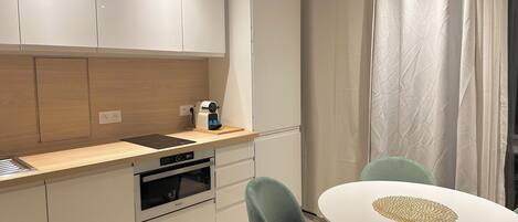 A modern kitchen with white cabinets, a built-in oven, and a coffee machine. There is a dining area with a round table and two chairs.
