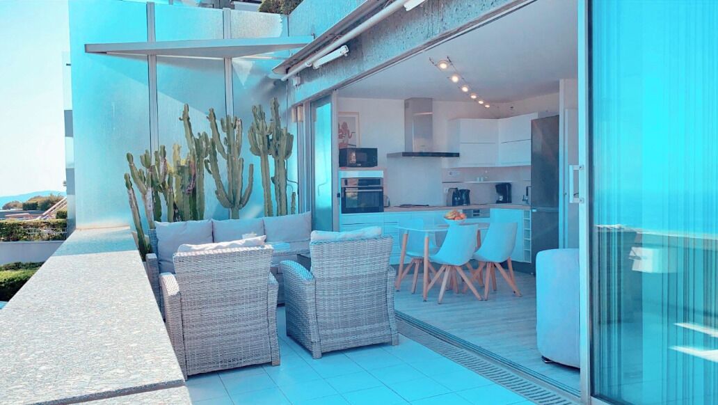 Un patio moderno al aire libre con mobiliario de mimbre, un ventanal de vidrio que da a una cocina y una planta de cactus.