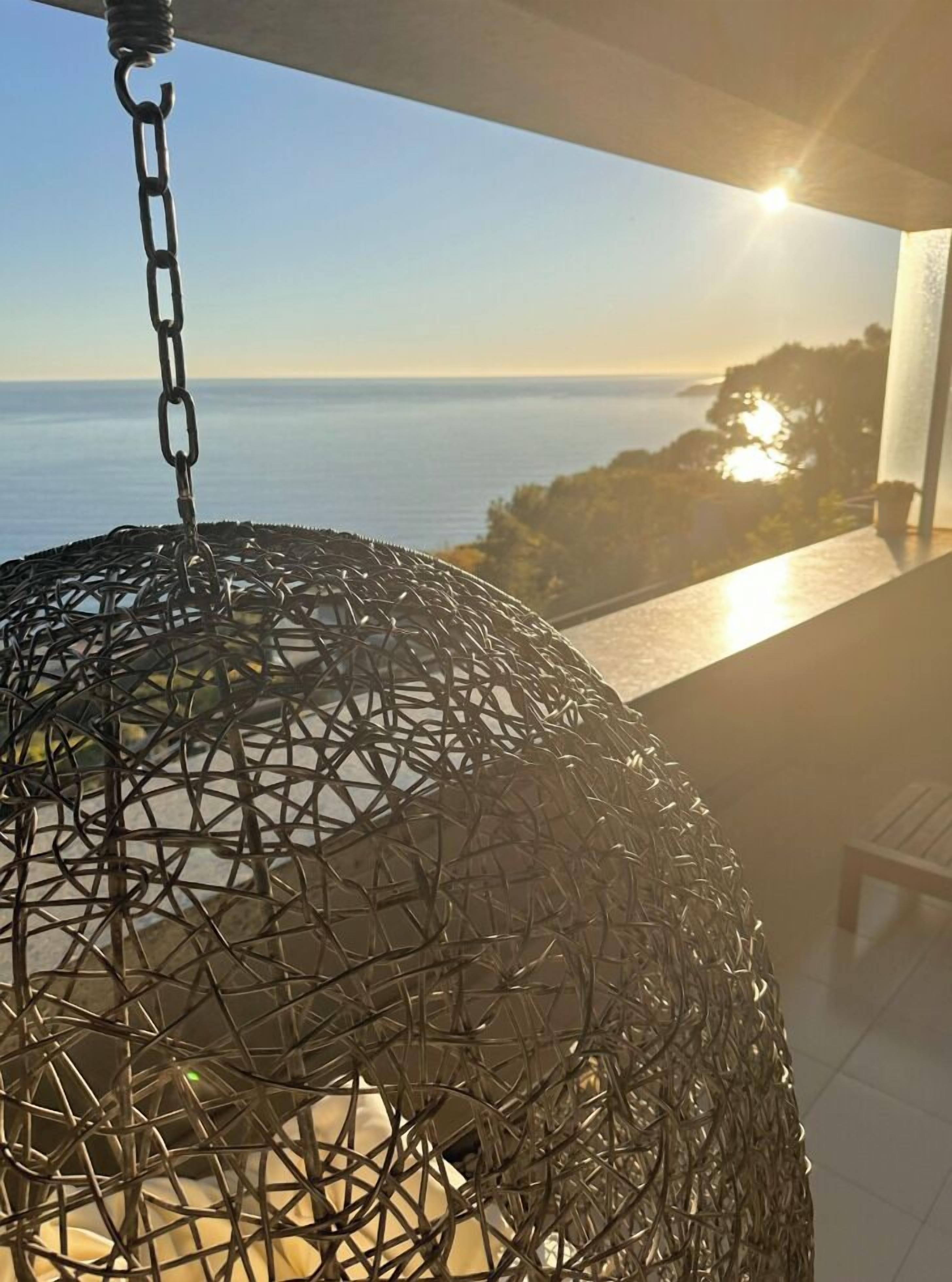 En hængende metalkugle med kæde, en balkon og udsigt til hav og træer.