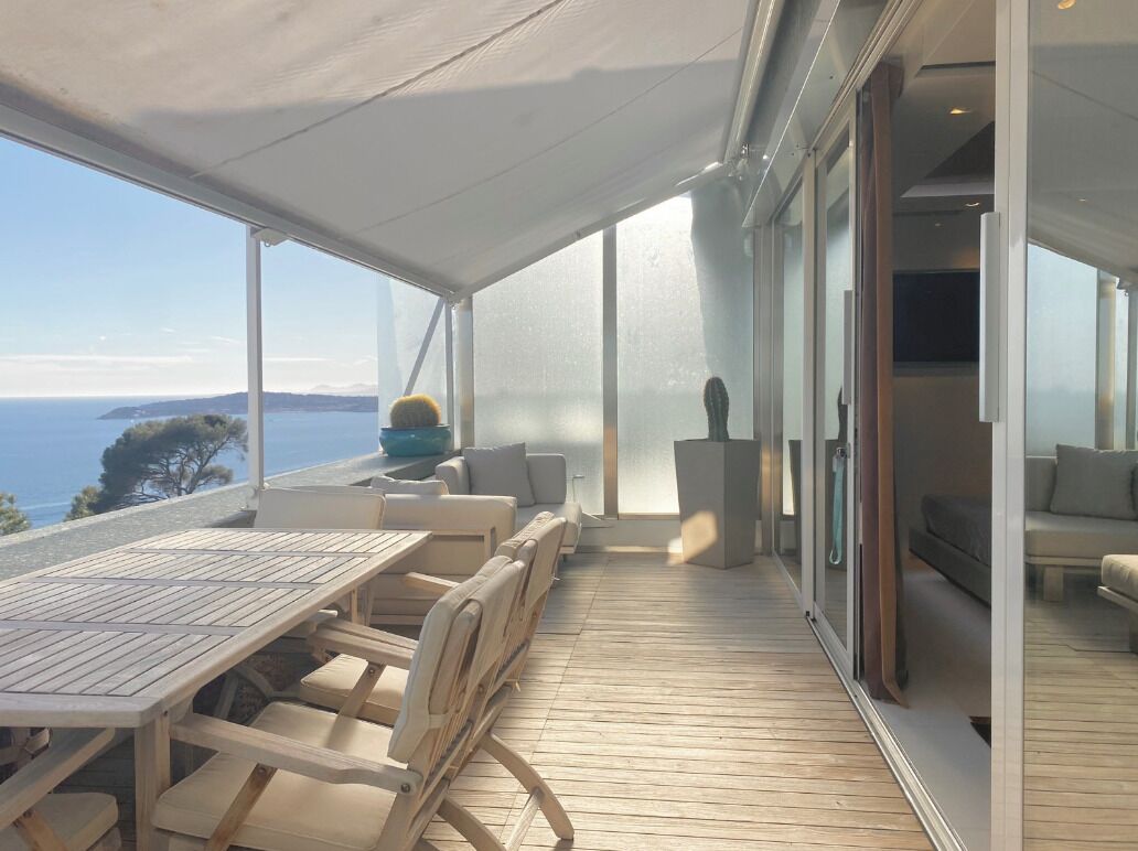A sunlit outdoor deck with a wooden table and chairs, a glass door leading to an interior space, and a view of the sea.