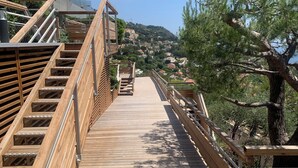 Une terrasse en bois avec des escaliers menant à un niveau supérieur, entourée d’arbres et offrant une vue sur une colline parsemée de bâtiments.